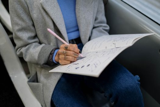 Personne assise dans un train, écrivant dans un carnet de stylisme avec un stylo rose. Vêtue d'un manteau gris et d'un pull b